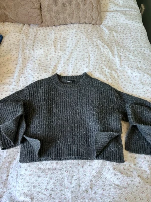 Grå stickad tröja från Gina Tricot - Mysig grå stickad tröja från Gina Tricot i storlek XS. Tröjan har en croppad passform, breda ärmar och ribbstickad struktur. Perfekt för lager-på-lager och passar till både jeans och kjol.