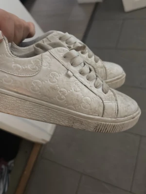 Vita sneakers med Mickey Mouse-mönster - Säljer ett par vita sneakers med präglat Mickey Mouse-mönster över hela skon. Skorna har klassisk låg profil, vita snören och sula i gummi. Materialet ser ut att vara konstläder och designen är clean men lekfull med Disney-detaljer.