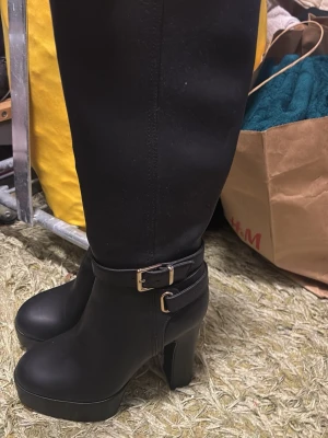 Svarta höga platåstövlar med spänne - Säljer ett par svarta höga stövlar med chunky platåklack och dekorativa spännen i guld vid ankeln. Stövlarna är i ett matt material och har en stilren, modern look som passar perfekt till höst- och vinteroutfits.