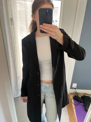 Svart trenchcoat med knappar - Säljer en svart trenchcoat med klassisk design och knappar framtill. Jackan har en rak passform, krage och bälte i midjan som ger en snygg siluett. Perfekt för dig som vill ha en stilren och tidlös look. Materialet känns mjukt och lätt.