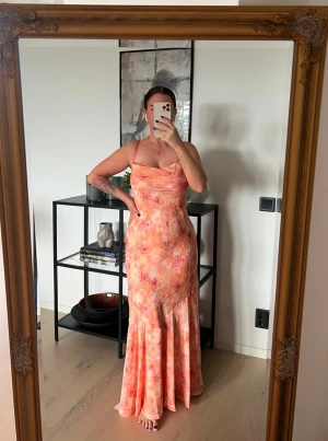 Adoore klänning - mer som strl 38 - Säljer en lång, figurnära klänning med smala axelband och blommigt mönster i orange, rosa och ljusa toner. Klänningen har en mjukt draperad urringning och är tillverkad i ett lätt, följsamt material som passar perfekt till sommaren.