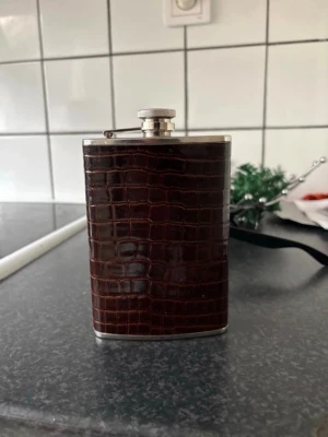 Mörkbrun fickplunta med krokodilmönster - Snygg fickplunta i rostfritt stål med mörkbrunt konstläder i krokodilmönster. Pluntan har en klassisk form och ett skruvlock med fäste så locket inte tappas bort. Perfekt att ta med på utflykten eller ge bort som present.