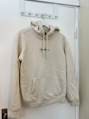 Beige hoodie från Hollister med logga - Snygg beige hoodie från Hollister med broderad svart logga på bröstet. Hoodien har en stor känguruficka framtill, dragsko i huvan och mjukt, fleeceliknande innerfoder. Perfekt för en avslappnad och trendig stil.