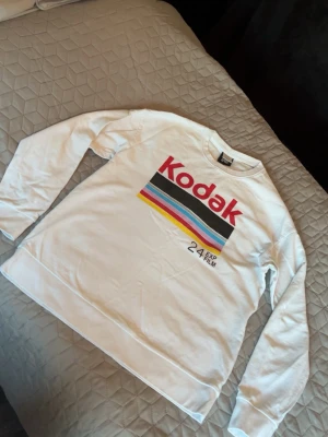 Vit Kodak sweatshirt med tryck - Säljer en vit sweatshirt från Kodak med färgglatt retrotryck och texten 'Kodak 24 EXP FILM' på bröstet. Tröjan har rund halsringning och långa ärmar. Perfekt för dig som gillar vintage och streetstyle. Storlek M
