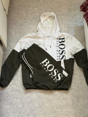 Hugo Boss  svart/vit - Säljer ett par snygga  från Hugo Boss i svart och vitt med stort BOSS-tryck på benet. Byxorna har resår i midjan och dragsko för skön passform. Matchar perfekt med hoodie i samma stil för en clean och modern look. Material som en regn jacka ! St S/xs lite smutsig här och där men ser ganska bra ut kan vara några brunna fläckar men syns knappast  material som en regn jacka