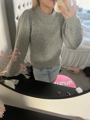 Grå stickad tröja med puffärm - Mysig grå stickad tröja med puffärmar och ribbade muddar vid ärmslut och hals. Tröjan har en rak passform och är perfekt för kyliga dagar. Materialet känns mjukt och värmande, och den neutrala färgen gör den enkel att matcha med olika outfits.