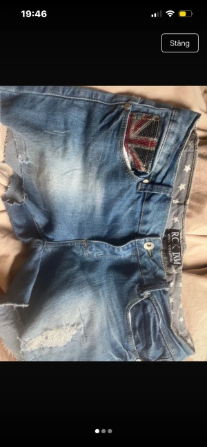 Blå jeansshorts från RCK DNM - Säljer ett par blå jeansshorts från RCK DNM med råa kanter och slitningar. Shortsen har en cool detalj med Union Jack-flagga i paljetter på fickan och stjärnmönstrat innerfoder. Perfekta för en avslappnad och trendig look. Säljer pga att de är för stora för mig och att jag behöver pengar! Strl s-m 