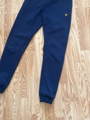 Mörkblå mjukisbyxor från Lyle & Scott - Mörkblå mjukisbyxor från Lyle & Scott med klassisk gul logga på höften. Byxorna har resår i midjan och vid bensluten samt dragkedjeficka. Perfekta för en avslappnad stil och tillverkade i mjukt material.