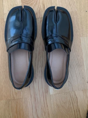 Svarta Tabis med delad tå. - Snygga svarta loafers i glansigt material med unik delad tå-design. Skorna har klassisk pennyloafers-look med markerad sula och stilren ovandel. Perfekta för dig som vill ha något annorlunda och trendigt i garderoben.
