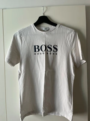 Vit Hugo Boss t-shirt med tryck - Snygg vit t-shirt från Hugo Boss med klassiskt svart logotryck på bröstet. T-shirten har rund halsringning och korta ärmar. Tillverkad i mjuk bomull för en skön känsla och enkel stil.