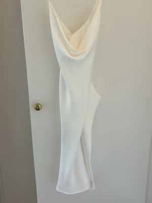 Vit klänning med draperad urringning - Elegant vit långklänning med draperad urringning och smala axelband. Klänningen har en slank siluett och är tillverkad i ett glansigt, mjukt material som ger en lyxig känsla. Perfekt för speciella tillfällen och stilrena outfits.