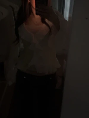 Vit volangblus från Nelly - Supersöt vit blus från Nelly med volangdetaljer längs v-ringningen och framtill. Blusen har ett vågigt, strukturerat tyg och långa ärmar med lätt utsvängda ärmslut. Perfekt att styla med jeans eller kjol för en trendig look.