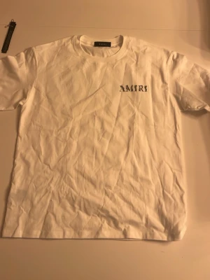 Vit t-shirt från Amiri - Säljer en clean vit t-shirt från Amiri med svart logga tryckt på bröstet. Klassisk rund hals och korta ärmar. Tillverkad i mjuk bomull som känns skön mot huden. Perfekt till jeans eller shorts för en enkel men snygg look. Perfekt för sommaren