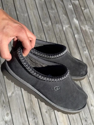 Gråa UGG Tasman - Säljer ett par mörkgrå UGG Tasman i mjuk mocka med broderad kant i svart och vitt. Insidan är fodrad med fluffigt material för extra komfort. Perfekta att glida runt i hemma eller utomhus när du vill ha det extra mysigt. Väldigt bra skick. Skriv vid frågor!