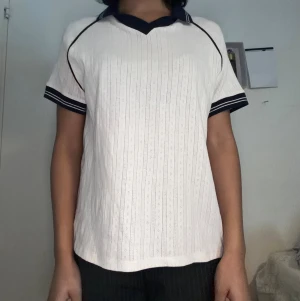 Vit t-shirt med marinblå krage - Vit t-shirt med marinblå detaljer och krage. T-shirten har korta ärmar och ett diskret hålmönster i tyget. Snygg kontrast mellan den vita basen och de mörkblå kanterna vid ärmslut och hals. Perfekt för en stilren och sportig look.