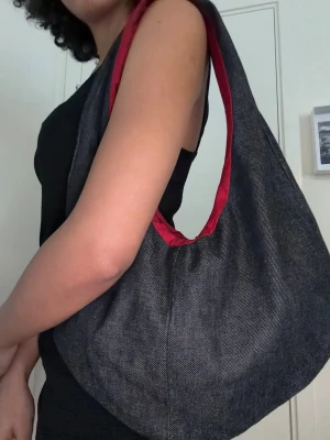 Svart denim väska - Stilren väska i svart denim med en snygg röd insida . Väskan har en mjuk, rundad form och bred axelrem som gör den bekväm att bära. Perfekt för dig som gillar en enkel men cool look.