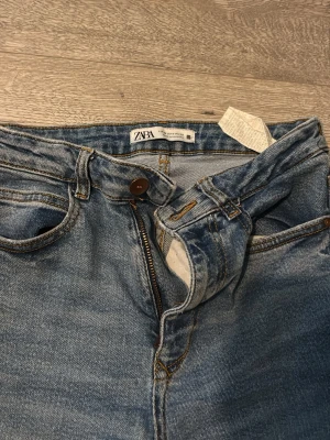 Blå utsvängda jeans från Zara - Säljer ett par blå jeans från Zara med utsvängda ben och klassisk femficksmodell. Jeansen har hög midja, dragkedja och knapp framtill samt snygga slitningar för en trendig look. Perfekta för dig som gillar retrostil och vill ha ett par bekväma jeans i bomull.