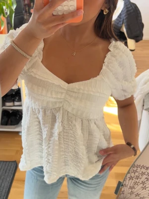 Vit puffärmad blus med struktur - Supersöt vit blus med puffärmar och smockad struktur. Blusen har en djupare urringning och volangdetaljer över bysten och ärmarna. Tillverkad i ett lätt, krinklat tyg som ger en luftig känsla. Perfekt att styla med jeans eller kjol för en trendig look. HELT NY