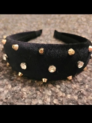 Svart diadem med nitar och strass - Snyggt svart diadem i sammet med guldiga nitar och glittrande strassdetaljer. Bred modell som ger en cool och trendig look, perfekt för att lyfta vilken outfit som helst. Materialet är mjukt och bekvämt mot håret.