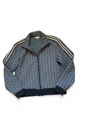 Lacoste monogram - Snygg blå track jacket från Lacoste med diskret mönster och klassiska ränder på axlarna. Dragkedja framtill, ribbade muddar och liten broderad krokodil-logga på bröstet. Tillverkad i polyester och bomull för en sportig känsla. Inga defekter helt ny köpt retail för 2,4!