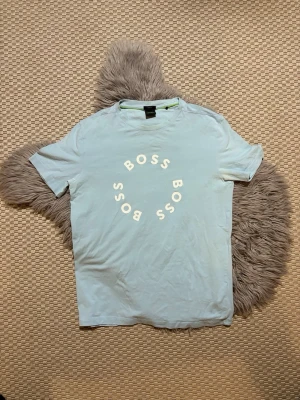 Ljusblå BOSS t-shirt med tryck - Snygg ljusblå t-shirt från BOSS med rund hals och vit cirkelformad logotyp på bröstet. T-shirten har en klassisk passform och är tillverkad i mjuk bomull, perfekt för en avslappnad och trendig look.