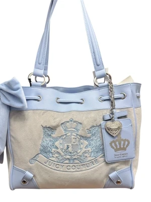 Juicy Couture ljusblå handväska - Superfin handväska från Juicy Couture i ljusblått och beige tyg med ljusblåa läderdetaljer. Väskan har broderad logga framtill, silverfärgade metalldetaljer, en söt rosett på sidan och en tag med hjärtberlock. Perfekt för dig som gillar en preppy och trendig look.
