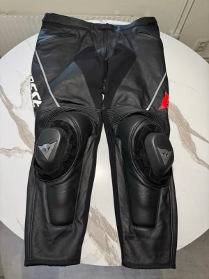 Svarta skinnbyxor från Dainese - Snygga svarta skinnbyxor från Dainese med vita och röda detaljer. Byxorna har inbyggda knäskydd och förstärkningar, perfekta för motorcykelåkning. Stretchpaneler för extra rörlighet. På insidan är storleken 28, motsvarar ca 52-54. Köp med eget ansvar.