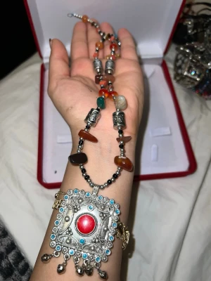 Bohemiskt halsband med färgglada pärlor - Unikt halsband med stora färgglada pärlor i rött, grönt, orange och svart. Halsbandet har ett stort, fyrkantigt hänge i silverfärgad metall med röda och turkosa detaljer samt små hängande dekorationer. Perfekt för dig som gillar bohemisk och statement-stil.