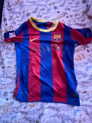FC Barcelona matchtröja Nike - Säljer en klassisk FC Barcelona matchtröja från Nike med röda och blå vertikala ränder, gul krage och klubbmärke på bröstet. Tröjan har korta ärmar, LFP-märke på ärmen och är tillverkad i lätt och ventilerande polyester. Perfekt för fotbollsträning eller att bära på läktaren.