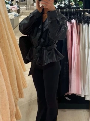 Svart skinnjacka med bälte från Zara - Snygg svart skinnjacka med markerade axlar och bälte i midjan som ger en cool siluett. Jackan har bred krage och en trendig, lite oversized passform. Perfekt för dig som vill ha en edgy look. Slutsåld