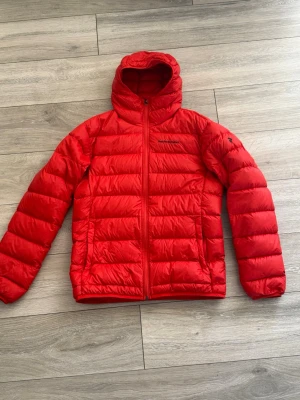 Röd dunjacka med huva från Peak Performance - Snygg röd dunjacka från Peak Performance med huva och dragkedja framtill. Jackan har två sidofickor, enkel och clean design med logga på bröstet. Perfekt för kalla vinterdagar och riktigt lätt att bära. Klassisk passform och stilren look. Pris kan diskuteras vid snabb affär 