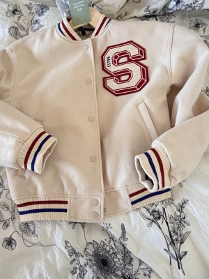 Beige bomberjacka med röd S-detalj - Stilren beige bomberjacka med broderad röd S och EST 96 på bröstet. Jackan har ribbade muddar och krage med röda och blå ränder, samt två snedställda fickor framtill. Stängs med tryckknappar. Perfekt för en sportig och trendig look. 