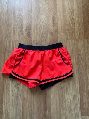 Orange Röda träningsshorts från Superdry - Snygga orange röda träningsshorts från Superdry med svarta detaljer och elastisk midja. Tillverkade i ett lätt och snabbtorkande material som transporterar bort fukt. Perfekta för löpning eller gymmet.