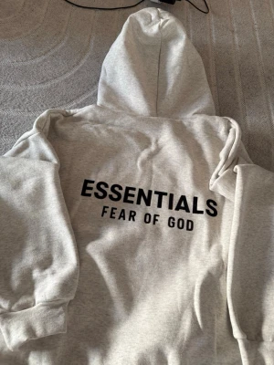Grå Essentials Fear of God hoodie - Snygg grå hoodie från Fear of God Essentials med svart logga tryckt på ryggen och mindre logga på bröstet. Oversized passform, mjukt material och stor huva. Perfekt för dig som gillar streetwear och vill ha en clean look.