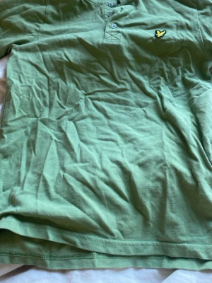 Grön pikétopp från Lyle & Scott - Säljer en grön pikétopp från Lyle & Scott Junior med klassisk krage och knappar framtill. Toppens material är mjuk bomull och den har ett broderat logomärke på bröstet. Perfekt för dig som gillar stilrena och sportiga plagg.