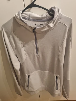 Ljusgrå hoodie från Under Armour - Snygg ljusgrå hoodie från Under Armour med halv dragkedja framtill och två fickor med dragkedja. Tillverkad i ett tekniskt funktionsmaterial som andas. Perfekt för träning eller chill. Huva och diskreta sömdetaljer för sportig look.