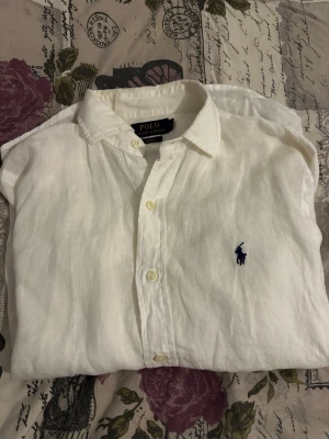 Vit linneskjorta Polo Ralph Lauren - Klassisk vit linneskjorta från Polo Ralph Lauren i slim fit-modell. Skjortan har knappar framtill, button down-krage och den ikoniska blå broderade loggan på bröstet. Perfekt för en clean och avslappnad stil.