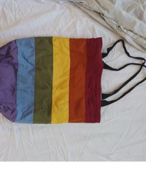 Pride tygväska - Säljer en färgglad shoppingväska i tyg med breda ränder i lila, blått, grönt, gult, orange och rött. Väskan har svarta axelband och är perfekt för att bära med sig till skolan, stan eller stranden. Enkel och stilren design som sticker ut.