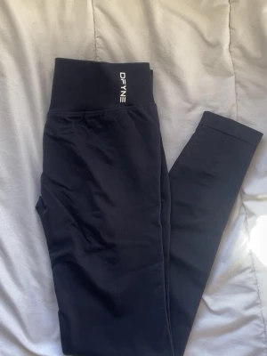 Svarta IMPACT leggings  DFYNE - Svarta seamless leggings från DFYNE med hög midja och ribbad linning. Materialet är stretchigt och följsamt, perfekt för träning eller chill. Diskret logga framtill och scrunch-detalj bak för snygg passform.