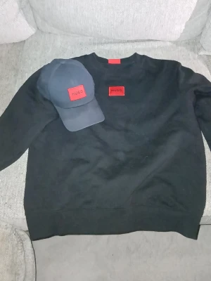 Svart sweatshirt från Hugo + matchande keps  - Svart sweatshirt från Hugo med röd logotyp-patch på bröstet. Klassisk rund halsringning och ribbade muddar vid ärmslut och nederkant. Perfekt för en stilren och enkel look. Materialet känns mjukt och bekvämt mot huden.