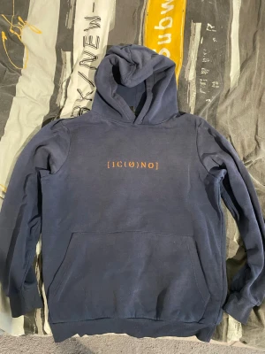Mörkblå hoodie från ICONO XS - Mörkblå hoodie från ICONO i regular fit med tryckt text '[IC(∅)NO]' på bröstet. Klassisk känguruficka framtill och ribbade muddar vid ärmslut och nederkant. Perfekt för en chill och avslappnad stil.