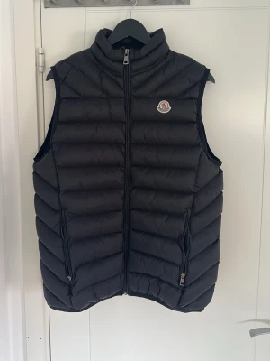 Svart dunväst från Moncler - Snygg svart dunväst från Moncler med hög krage och dragkedja framtill. Västen har två sidofickor med dragkedja och Monclers klassiska logotyp på bröstet. Tillverkad i glansigt material med quiltad design som ger en modern look.