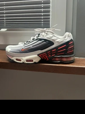 Nike Air Max Plus 3 vita och svarta - Nike Air Max Plus 3 sneakers i vitt, svart och rött med futuristisk design och synlig Air Max-dämpning. Ovandel i mesh och syntet, röda detaljer på sidorna och liten röd swoosh. Skorna har en chunky sula och coola linjer längs ovandelen.