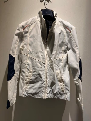 Vit vindjacka från BMW Sauber F1 - Snygg vit vindjacka från  BMW Sauber F1 med marinblå detaljer på ärmarna och diskreta röda inslag vid axlarna. Jackan har hög krage, dragkedja framtill och två fickor med dragkedja. Perfekt för dig som gillar sportig och stilren look.