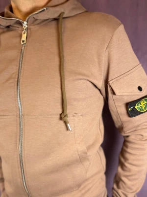 Beige hoodie från Stone Island - Säljer en beige hoodie från Stone Island med dragkedja och huva. Hoodien har en ikonisk patch på ärmen och snörning i huvan. Materialet är mjukt och bekvämt, perfekt för en avslappnad look. Fickor framtill och coola detaljer på ärmen.