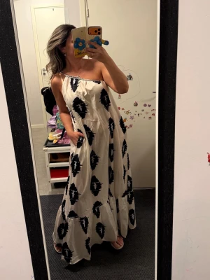 Vit långklänning med svart mönster - Säljer en vit långklänning med svart grafiskt mönster. Klänningen har axelbandslös design och kan stylas med eller utan bälte i midjan. Bälte EJ ingår.Luftigt material, perfekt för varma dagar. Snygg och trendig look som sticker ut.helt ny