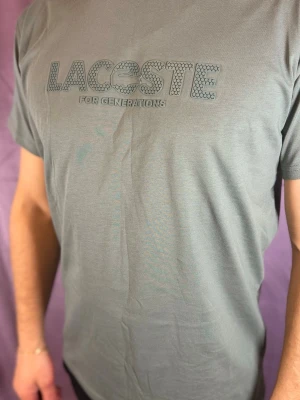 Grå t-shirt från Lacoste - Säljer en grå t-shirt från Lacoste med coolt tryck på bröstet där det står 'Lacoste for generations'. T-shirten har korta ärmar och är gjord i mjuk bomull, perfekt för dig som gillar stilrena plagg med en twist.