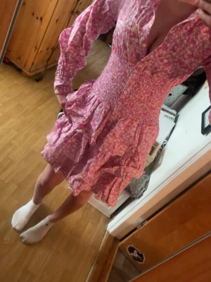 Rosa blommig kortklänning med volang - Supersöt rosa kortklänning med blommigt mönster och volangdetaljer. Klänningen har långärmade ärmar, markerad midja och v-ringning. Perfekt för vår och sommar, och ger en riktigt fräsch och ungdomlig vibe.