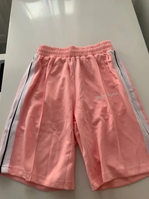 Palm Angels rosa shorts med ränder - Snygga rosa shorts från Palm Angels med vita och svarta ränder längs sidorna. Shortsen har elastisk midja och logga broderad på vänster sida. Perfekta för en avslappnad och trendig look.