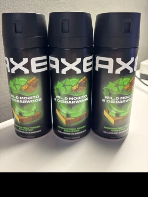 Axe Wild Mojito & Cedarwood Body Spray - Tre svarta flaskor Axe body spray med doften Wild Mojito & Cedarwood. Flaskorna har en grön och brun etikett med motiv av en mojito och cederträ. Sprayen är designad för att ge en fräsch och uppfriskande känsla hela dagen.